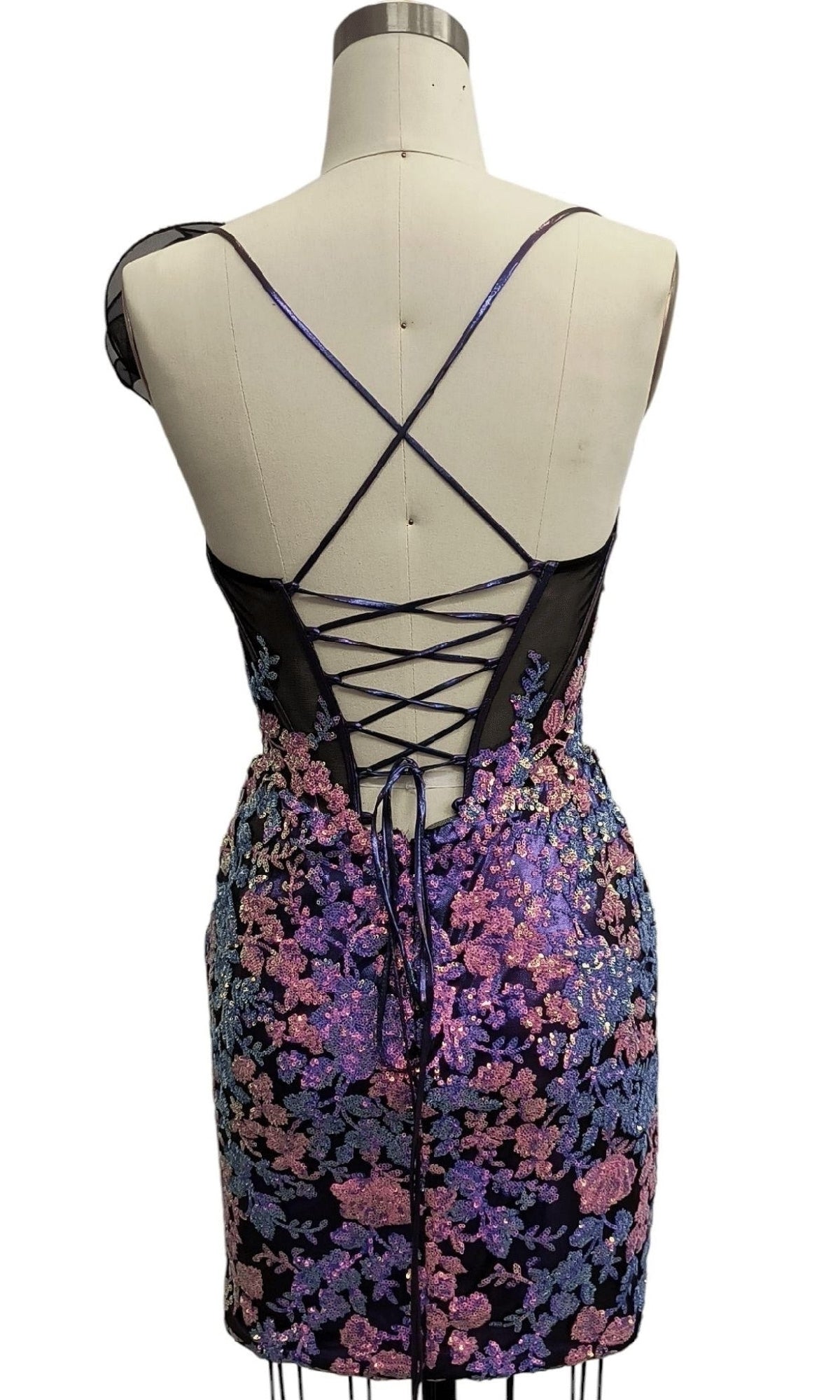 A mannequin displays the Short Purple Sequin Hoco Dress 12567, a sleeveless, short designer dress with a crisscross lace-up back and floral sequin embroidery in purple tones.